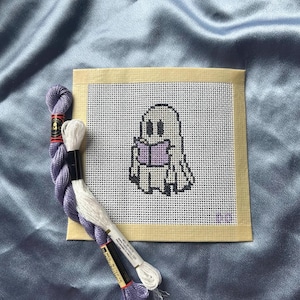 May include: A square needlepoint canvas featuring a pixelated ghost character reading a purple book, outlined in black thread. Two skeins of embroidery floss, one lavender and one white, are positioned next to the canvas on a blue fabric.