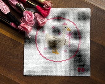 May include: A square needlepoint canvas featuring a chicken design within a pink circle. The chicken is yellow and pink, with pink accents. Pink embroidery floss is arranged around the canvas, ready for stitching. The canvas has the number "00" in the bottom right corner.