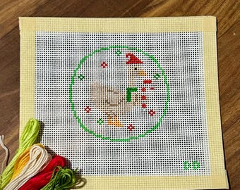 May include: A needlepoint canvas featuring a whimsical design of a snowman wearing a red hat and scarf, encircled by a green border. The canvas is surrounded by a yellow border and accompanied by colorful embroidery floss in shades of green, red, yellow, and cream.
