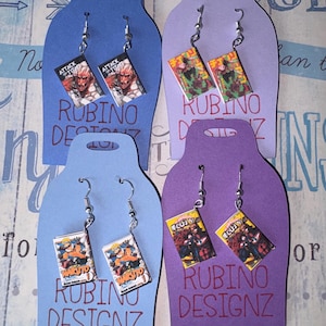 May include: Four pairs of dangle earrings featuring miniature manga book covers. The designs include Attack on Titan, My Hero Academia, and Naruto. Each pair is attached to a colored cardstock display tag with the text RUBINO DESIGNZ printed.