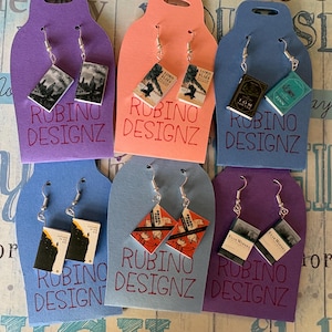 May include: A collection of book-themed earrings displayed on colorful cardstock. Each earring set features miniature book covers with titles like "Lord of the Flies" and "Tom Sawyer." The cardstock has the text "RUBINO DESIGNZ" in red.