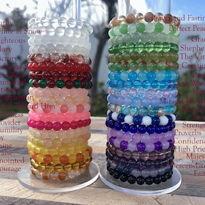May include: Two stacks of beaded bracelets in various colors, including clear, red, pink, yellow, green, blue, and purple. Each bracelet is made of round beads. Text on the side lists words like "Purity", "Victory", and "Strength".