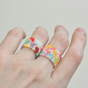 May include: Two colourful beaded rings are shown on a hand. The rings feature a variety of colours, including red, yellow, blue, and green. The rings are made of small beads and have a textured appearance. The rings are a unique and eye-catching accessory.