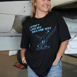 Puede incluir: Camiseta negra con el texto "I DON'T LIKE MY BEAVER WET" en azul claro. El gráfico representa un hidroavión. La camiseta se combina con unos vaqueros azules. El fondo incluye una parte de un avión.