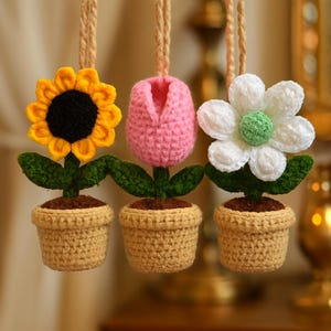 Puede incluir: Tres adornos florales de ganchillo: un girasol amarillo con centro negro, un tulipán rosa y una margarita blanca con centro verde. Cada flor está en una maceta de ganchillo beige con 'tierra' marrón y hojas verdes.