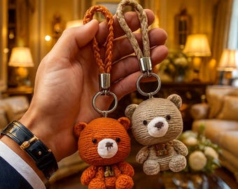 May include: Two handmade teddy bear keyrings. One is orange, the other is tan. Both have white accents on their faces and small decorative elements. The keyrings have braided leather straps and silver-coloured metal rings.