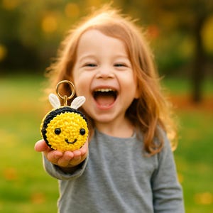 May include: A yellow and black crocheted bee keychain with white wings and a gold-colored keyring. The bee has a smiling face with black eyes. The keychain is held up by a person with long red hair.
