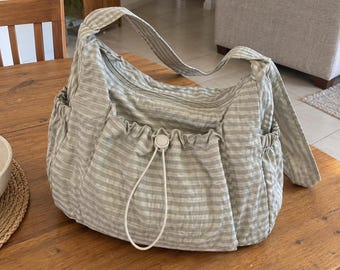 Könnte beinhalten: Eine weiche, lässige Schultertasche im Hobo-Stil mit einem hellgrau-weißen horizontalen Streifenmuster. Die Tasche verfügt über einen Reißverschluss oben, einen breiten Schulterriemen und eine Außentasche vorne mit gerafftem Rand und weißem Kordelzug.