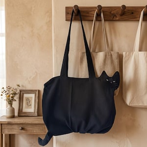 K&ouml;nnte beinhalten: Eine schwarze Tragetasche aus Segeltuch mit einem katzenf&ouml;rmigen Design, spitzen Ohren, wei&szlig; gestickten Augen und einem langen Schwanz, der aus der unteren Ecke ragt. Die Tasche hat zwei lange Schulterriemen und h&auml;ngt an einem Holzhaken.