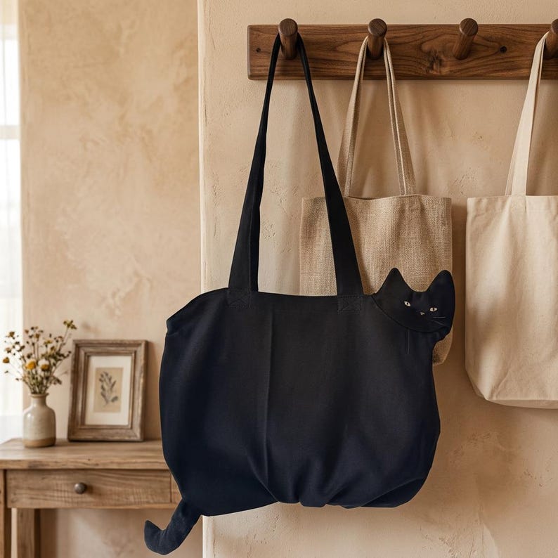K&ouml;nnte beinhalten: Eine schwarze Tragetasche aus Segeltuch mit einem katzenf&ouml;rmigen Design, spitzen Ohren, wei&szlig; gestickten Augen und einem langen Schwanz, der aus der unteren Ecke ragt. Die Tasche hat zwei lange Schulterriemen und h&auml;ngt an einem Holzhaken.