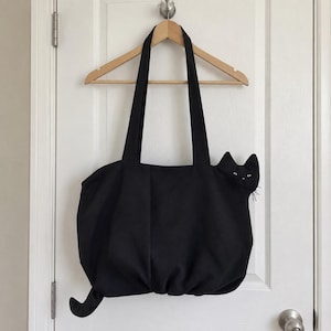 K&ouml;nnte beinhalten: Eine schwarze Tragetasche mit einem katzenf&ouml;rmigen Design, bei dem ein Kopf an der rechten Seite hervorsteht, komplett mit gestickten Augen und Schnurrhaaren, sowie einem geschwungenen Schwanz an der unteren linken Ecke, an zwei langen Tr&auml;gern.