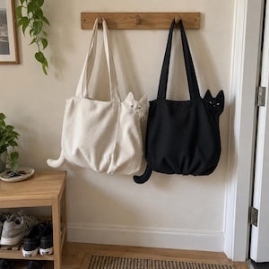 K&ouml;nnte beinhalten: Zwei Stofftaschen h&auml;ngen an einer Holzhakenleiste. Eine Tasche ist cremefarben, die andere schwarz. Jede Tasche hat einen dreidimensionalen Katzenkopf an der Seite mit Schnurrhaaren und Ohren sowie einen Katzenschwanz an der unteren Ecke.