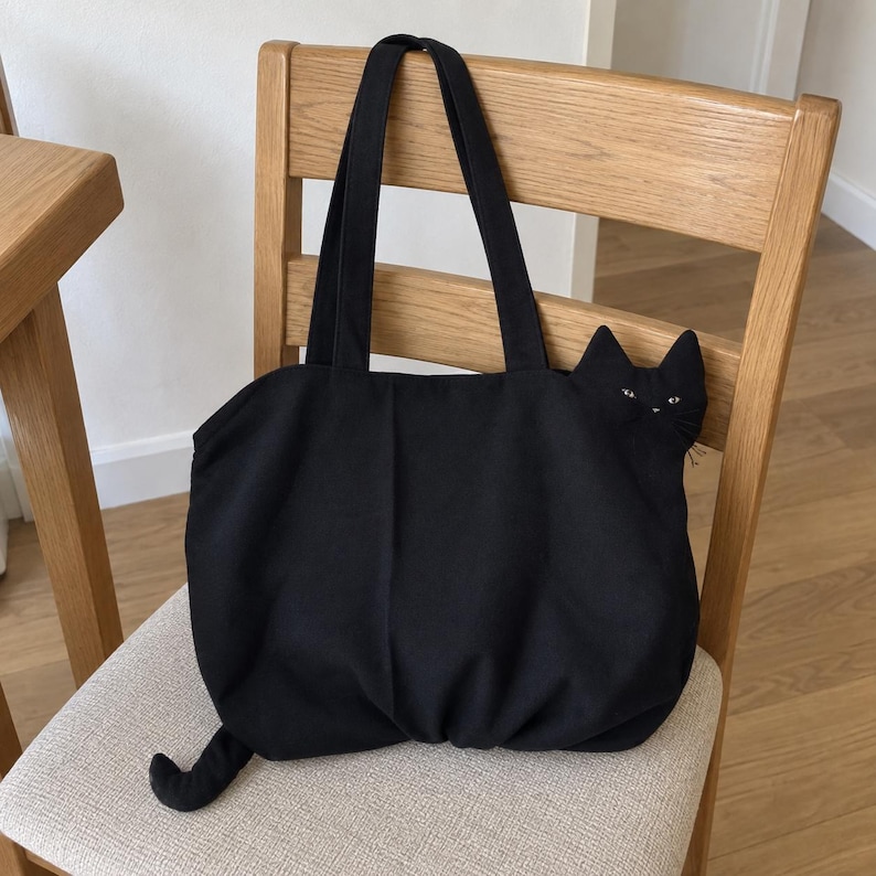 K&ouml;nnte beinhalten: Eine schwarze Tragetasche mit einem dreidimensionalen Katzenkopf an der oberen rechten Seite und einem kleinen Katzenschwanz, der aus der unteren linken Ecke ragt. Die Tasche hat zwei lange Schulterriemen und eine weiche, geraffte Stoffstruktur.