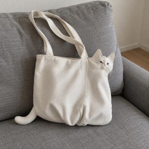 K&ouml;nnte beinhalten: Eine cremefarbene Canvas-Tragetasche mit einem dreidimensionalen Katzenkopf an der rechten Seite und einem Katzenschwanz, der unten links hervorsteht. Die Tasche hat zwei lange Schulterriemen und eine strukturierte Stoffoberfl&auml;che mit Katzendesign.