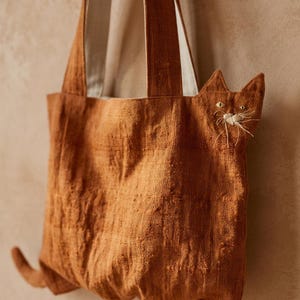 K&ouml;nnte beinhalten: Eine rostfarbene Tragetasche mit strukturierter Stoffoberfl&auml;che, die an der Seite eine integrierte katzenf&ouml;rmige Silhouette aufweist. Das Katzendesign umfasst gestickte Augen und wei&szlig;e Faden-Schnurrhaare. Die Tasche hat zwei Trageriemen und einen kleinen Schwanz.