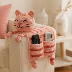 May include: A crocheted cat-shaped armrest organizer in light pink with darker pink stripes. The cat head sits on top, while the body drapes over the side, featuring two pockets holding a black remote control and a white digital remote device.