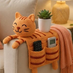 May include: An orange and brown striped crochet cat armrest organizer featuring a smiling cat head, two front pockets containing a black remote and a gray remote, and two dangling cat paws, with a small green succulent in a white pot on top.