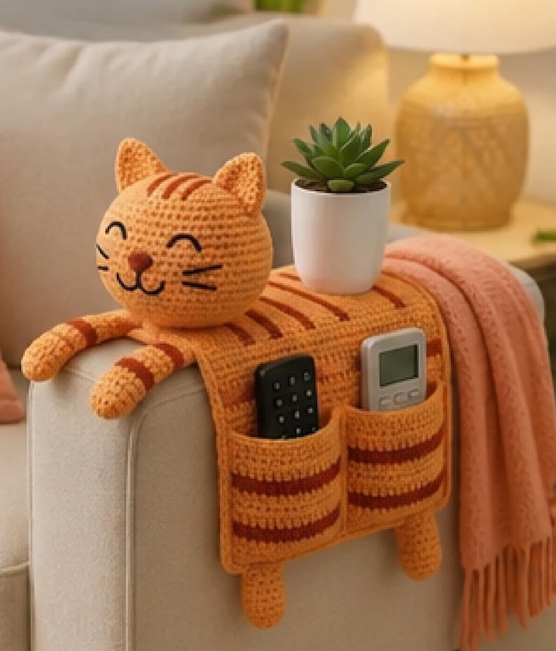 May include: An orange and brown striped crochet cat armrest organizer featuring a smiling cat head, two front pockets containing a black remote and a gray remote, and two dangling cat paws, with a small green succulent in a white pot on top.