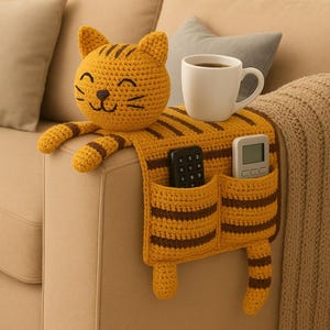 May include: A mustard yellow crochet sofa armrest organizer shaped like a cat with dark brown stripes. It features a cat head, two front paws, a tail, and two pockets holding a black remote control and a white digital remote. A white mug sits on top.