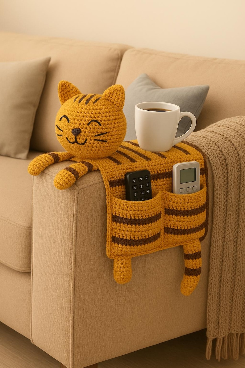 May include: A mustard yellow crochet sofa armrest organizer shaped like a cat with dark brown stripes. It features a cat head, two front paws, a tail, and two pockets holding a black remote control and a white digital remote. A white mug sits on top.