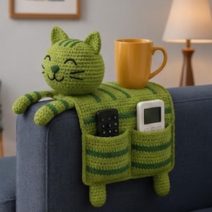 May include: A green crocheted cat-shaped sofa armrest organizer with dark green stripes. The cat head features embroidered facial details. The organizer includes two pockets holding a black remote and a white remote, with a yellow mug placed on top.