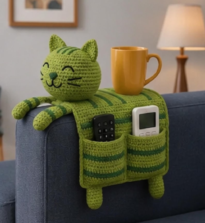 May include: A green crocheted cat-shaped sofa armrest organizer with dark green stripes. The cat head features embroidered facial details. The organizer includes two pockets holding a black remote and a white remote, with a yellow mug placed on top.