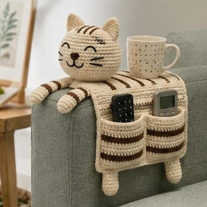 May include: A beige and brown striped crochet sofa armrest organizer shaped like a cat. The organizer features a cat head, two front paws, and two pockets containing a black remote control and a grey remote control. A white mug with black dots sits on top.