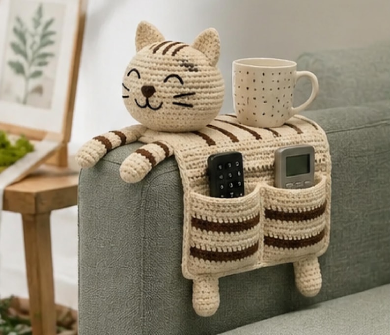 May include: A beige and brown striped crochet sofa armrest organizer shaped like a cat. The organizer features a cat head, two front paws, and two pockets containing a black remote control and a grey remote control. A white mug with black dots sits on top.
