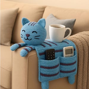 May include: A light blue crochet cat armrest organizer with dark purple stripes. The cat head features embroidered facial details. The body drapes over the armrest, featuring two pockets holding a black remote and a white remote, with a white mug on top.
