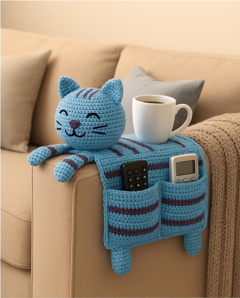 May include: A light blue crochet cat armrest organizer with dark purple stripes. The cat head features embroidered facial details. The body drapes over the armrest, featuring two pockets holding a black remote and a white remote, with a white mug on top.