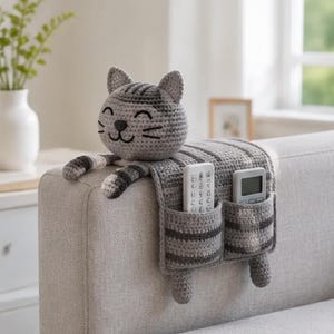 May include: A gray and dark gray striped crochet cat organizer draped over a sofa arm. The cat head features embroidered eyes and whiskers. Two rectangular pockets on the hanging panel hold a white remote control and a gray digital remote control.