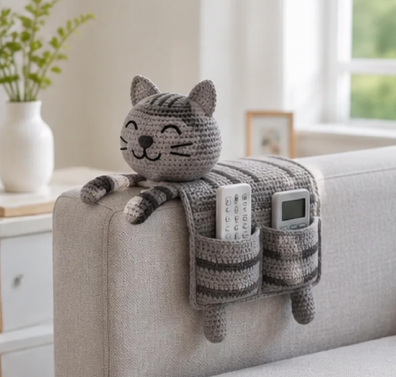May include: A gray and dark gray striped crochet cat organizer draped over a sofa arm. The cat head features embroidered eyes and whiskers. Two rectangular pockets on the hanging panel hold a white remote control and a gray digital remote control.