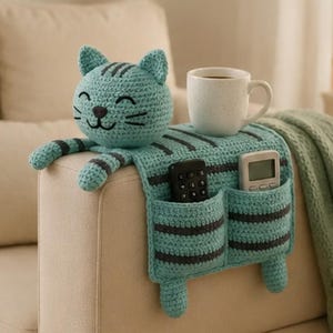 May include: A turquoise and dark gray striped crochet cat-shaped sofa armrest organizer featuring a head with embroidered facial features, two front paws, and two storage pockets containing a black remote control and a white digital device.