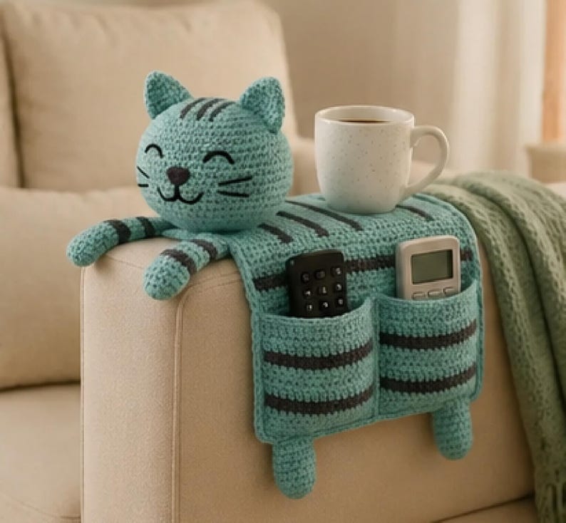 May include: A turquoise and dark gray striped crochet cat-shaped sofa armrest organizer featuring a head with embroidered facial features, two front paws, and two storage pockets containing a black remote control and a white digital device.