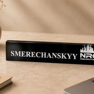 May include: A black desk nameplate with white lettering that reads "SMERECHANSKYY" and the NRG Noble Realty Group logo. The nameplate is rectangular and sits on a light-colored desk. A laptop, notebook, and pen are also visible.