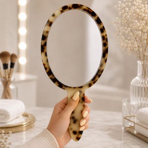 May include: Oval-shaped hand mirror with a tortoiseshell pattern frame and handle. The mirror is held up in front of a blurred background with makeup brushes, a vase of flowers, and a white towel. The mirror is a beauty accessory.