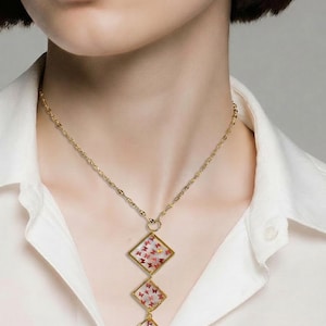May include: A gold-colored necklace with three diamond-shaped pendants. Each pendant contains small red and gold accents. The necklace is worn over a white shirt.