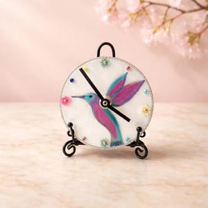 May include: A decorative clock featuring a hummingbird design. The clock face is white with a colorful hummingbird illustration in shades of blue, purple, and pink. The clock is adorned with small, colorful embellishments and set on a black metal stand.