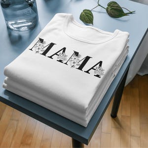 May include: A stack of white t-shirts featuring the word "MAMA" in black, decorated with floral designs. The shirts are neatly folded on a blue surface. A glass vase and greenery are visible in the background.