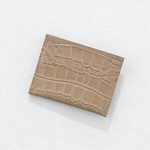 May include: A tan card holder with a crocodile skin texture. The rectangular wallet has multiple card slots and is stitched with a lighter tan thread. The card holder is made of a smooth, shiny material.