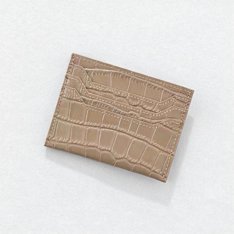 May include: A tan card holder with a crocodile skin texture. The rectangular wallet has multiple card slots and is stitched with a lighter tan thread. The card holder is made of a smooth, shiny material.