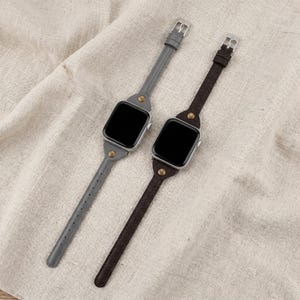 May include: Two smart watch bands, one gray and one brown, with silver buckles and gold-colored rivets. The bands are made of leather and are displayed on a neutral-colored fabric.