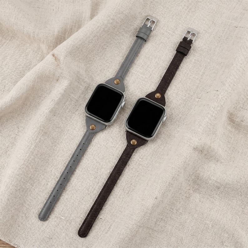 May include: Two smart watch bands, one gray and one brown, with silver buckles and gold-colored rivets. The bands are made of leather and are displayed on a neutral-colored fabric.