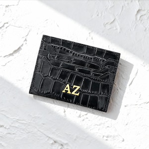 May include: Black card holder with a crocodile skin texture. The card holder has multiple slots and features the gold letters "AZ" on the front. The card holder is rectangular and sits on a white textured surface.