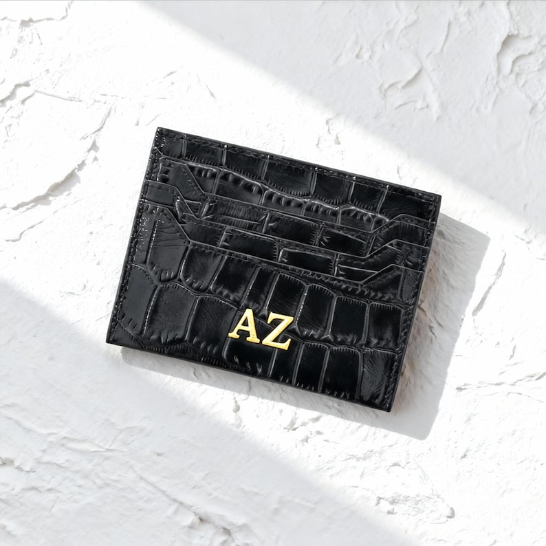 May include: Black card holder with a crocodile skin texture. The card holder has multiple slots and features the gold letters "AZ" on the front. The card holder is rectangular and sits on a white textured surface.