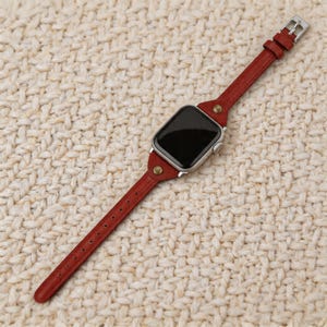 May include: A red leather watch band with a silver watch face. The band is attached with silver hardware and brass rivets. The watch band is shown on a textured, off-white surface.