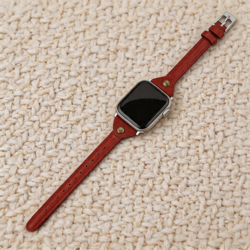 May include: A red leather watch band with a silver watch face. The band is attached with silver hardware and brass rivets. The watch band is shown on a textured, off-white surface.