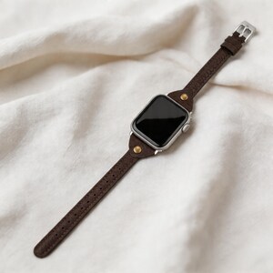 May include: A brown leather watch band with a silver watch face. The band is secured with a silver buckle and features decorative brass rivets. The watch face is black.