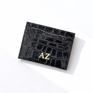 May include: Black card holder with a crocodile texture and the gold letters "AZ". The card holder has multiple slots for cards. The card holder is rectangular and made of a shiny material.