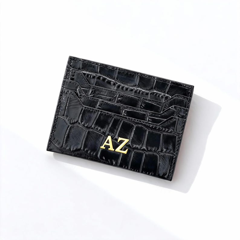 May include: Black card holder with a crocodile texture and the gold letters "AZ". The card holder has multiple slots for cards. The card holder is rectangular and made of a shiny material.